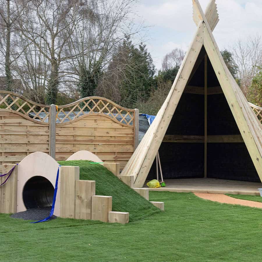 Wooden outdoor play area with a climbing tunnel and a teepee, designed for preschool children at Monkey Puzzle Day Nursery & Preschool Lightwater. Perfect for imaginative play and outdoor activities.